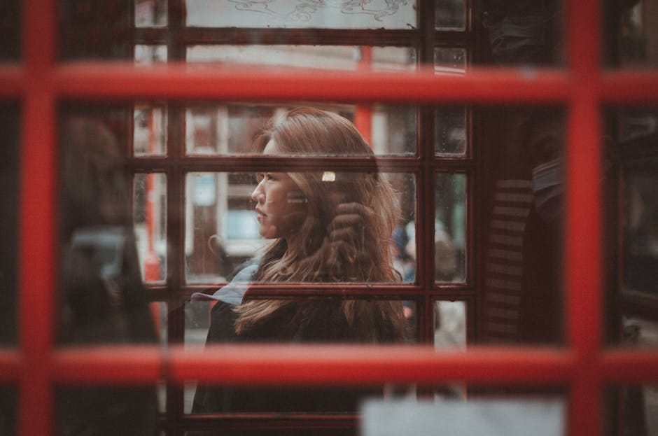 A woman gazes outside from inside a classic red London phone booth, capturing urban reflections.